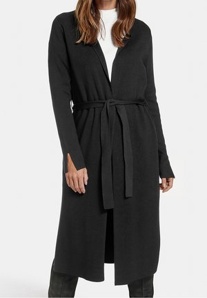 Femme portant un long cardigan noir ceinturé par-dessus un haut blanc et un pantalon noir, se tenant devant un fond clair uni.