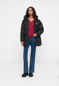 Žena u crnoj puffer jakni, bordo V-izrezani pulover, plavim trapericama i crnim čizmama stoji protiv bijele pozadine.