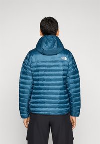Donkerblauwe pufferjack met horizontaal gestikte secties, een capuchon en een wit logo op de bovenrug. Glad, glanzend materiaal met isolatie.
