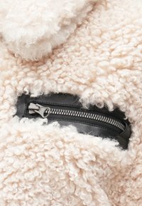 Prim-plan al unui material de fâșie curlicie de culoare crem, cu un buzunar cu fermoar negru, dotat cu o manetă robustă din metal.