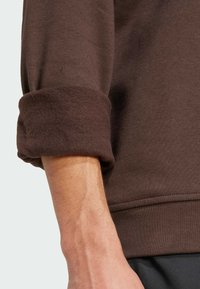 Hnědá látka svetru s texturovaným fleecem na manžetě. Detailní záběr ruky ukazující tón pleti a detaily zápěstí.