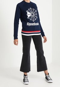 Námořnická modrá mikina s bílým geometrickým logem a textem "Reebok". Spárována s černými zvonovými džíny a černo-bílými teniskami.