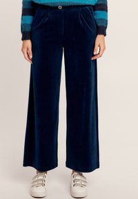 Marineblauwe corduroy flared broek met voorplooien, zijzakken en een knoopsluiting, gecombineerd met witte sneakers met klittenbandsluitingen.