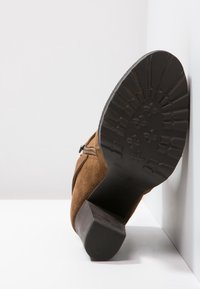 Brune ruskind ankelstøvle med en stablet hæl, har en tekstureret gummisål, lynlås i siden og rund tå-design.