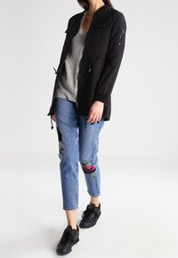 Schwarze Kapuzenjacke mit zwei Fronttaschen und einer Reißverschlusstasche am Ärmel; kombiniert mit blauen Jeans mit floralen Stickereien und schwarzen Sneakers.