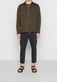 Giacca button-up verde oliva con tasche, indossata sopra una camicia scura, abbinata a pantaloni scuri a sigaretta e sandali neri.