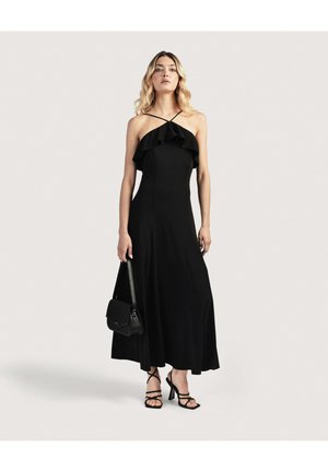 Naine kannab musta varrukateta maksikleit rüüsidega, hoiab käes musta käekotti ja tal on jalas mustad kontsaga rihmikud sandaalid.