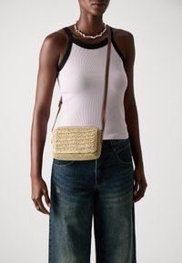 Small woven crossbody bag in natural straw color, rectangular shape with a textured surface, brown leather strap, and a flap closure.