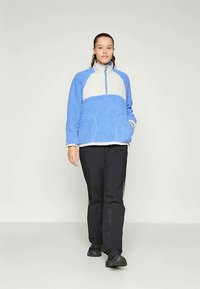 Giovane che cammina, indossando una giacca in fleece blu e bianca, pantaloni neri con strisce laterali e sneaker nere su uno sfondo semplice.