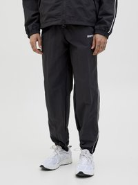 Svart nylon treningsdress med elastiske ermer, med hvite sidestriper og en liten logo. Båret med hvite treningssko.
