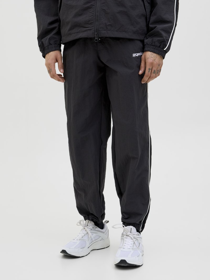 Svart nylon treningsdress med elastiske ermer, med hvite sidestriper og en liten logo. Båret med hvite treningssko.
