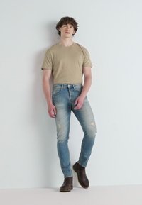 Bēša īsām piedurknēm t-krekls, izbalējuši zili skinny džinsi ar noberztām vietām un brūnas potītes zābaki. Modelis stāv pretim vienkāršai fona.