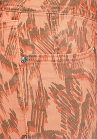 Persikofärgad tyg med ett brunt abstrakt mönster. Har fickor och silverdetaljer. Texturen verkar mjuk och liknar denim.