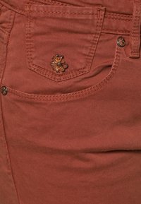Spodnie w kolorze rdzy z małą kieszenią ozdobioną kwiatowym haftem i kontrastowym przeszyciem. Gładka faktura materiału.