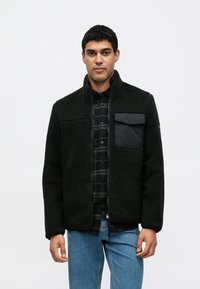 Fekete polárdzseki cipzáras előlappal, magas gallérral és egy fekete textil mellzsebbel. Fekete kockás inggel és kék farmerral párosítva.