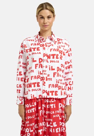 Frau mit blondem Haar, die ein weißes Hemd mit Knöpfen und einen roten Rock trägt, beide bedeckt mit roten und weißen Blockbuchstaben in Italienisch.