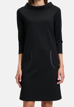 Femme portant une robe noire longueur genou avec des manches trois-quarts, un large décolleté et deux grandes poches avant avec bordure.