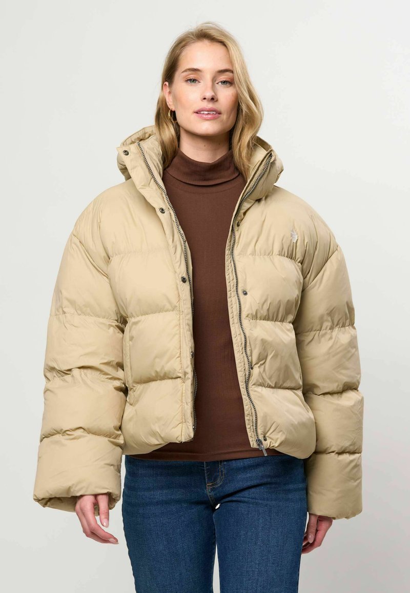 Femme portant une veste matelassée beige sur un col roulé marron et un jean bleu, debout devant un fond clair uni.