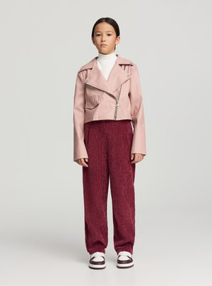 Giacca di pelle rosa corta con chiusura a zip, due tasche frontali, abbinata a pantaloni rossi scuri testurizzati e sneakers bianche.