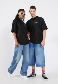 Black oversized t-shirts with a logo, paired with light blue denim pants. One model wears white sunglasses; the other wears black and white sneakers.