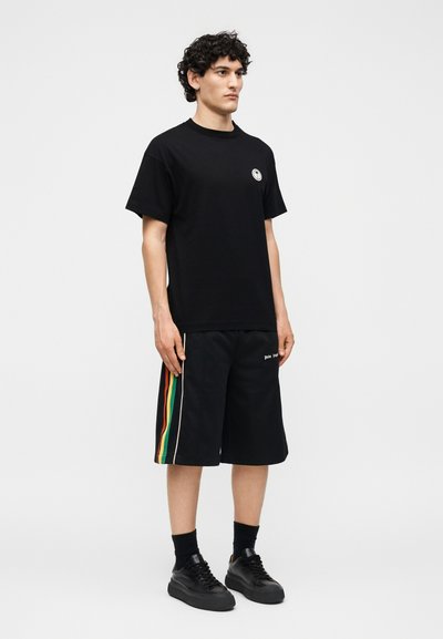 Άνδρας με σγουρά μαλλιά που φοράει μαύρο μπλουζάκι και μαύρο σορτς με κάθετες κόκκινες, κίτρινες και πράσινες ρίγες, στέκεται μπροστά σε μια απλή φόντο.