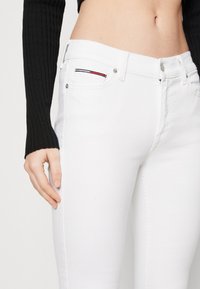 Vaqueros de mezclilla blanca con un diseño ajustado, que presentan un pequeño detalle de costura multicolor en el bolsillo frontal y herrajes plateados.