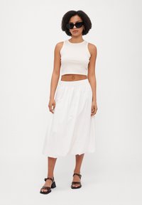 Bijeli crop top i duga, lepršava bijela suknja. Ravne sandale s kvadratnim vrhom i niskim blok petama. Dodatci uključuju velike crne sunčane naočale.