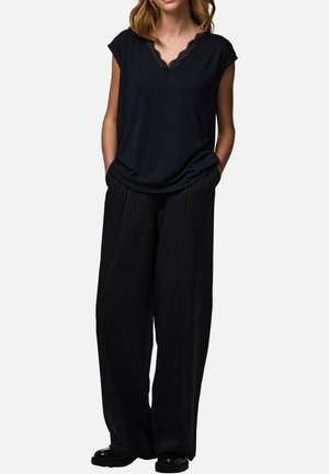 Kobieta w czarnym topie bez rękawów z koronkowym dekoltem w kształcie litery V oraz czarnych, szerokich spodniach w prążki, ręce w kieszeniach, stojąca na białym tle.