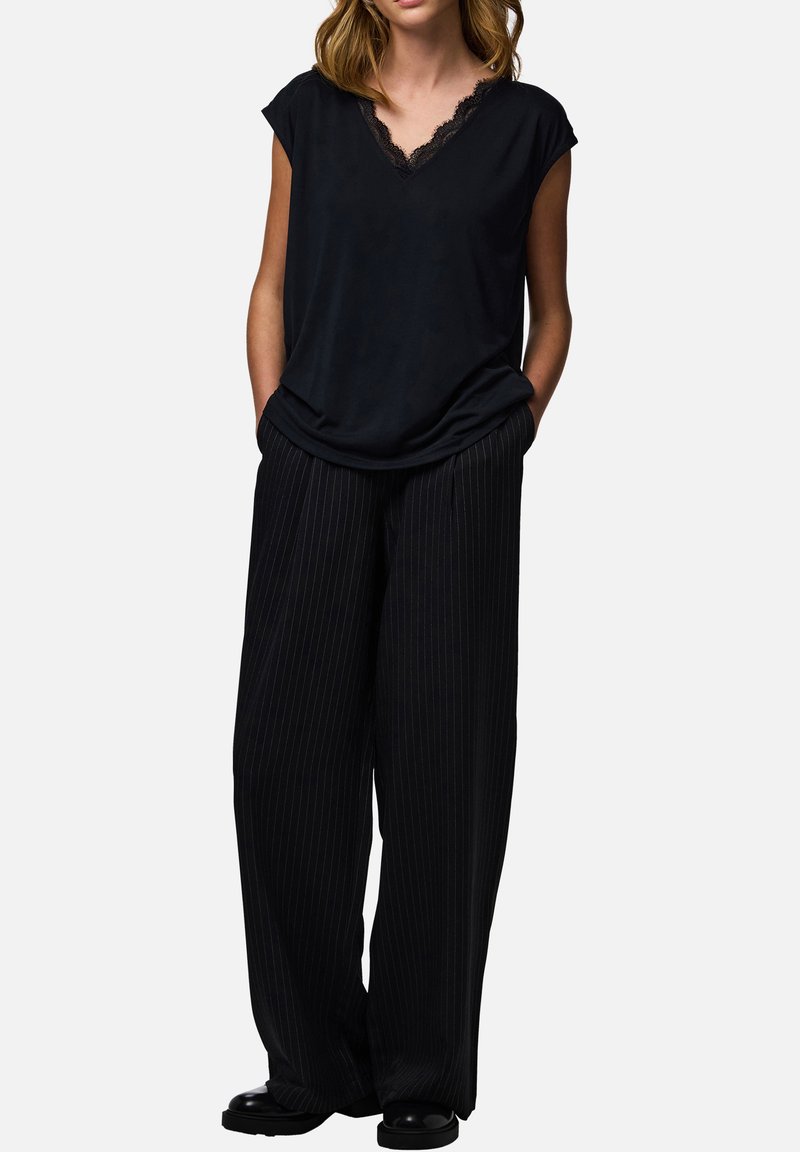 Femme portant un top noir sans manches avec un col en V en dentelle et un pantalon noir à jambes larges à rayures fines, les mains dans les poches, debout sur fond blanc.