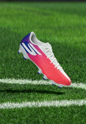 Chaussure de football aux couleurs rose, blanche et bleue flottant au-dessus d'un terrain herbeux près des lignes blanches de délimitation.