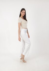 Joven mujer con top de mangas cortas beige, pantalones blancos de cintura alta y sandalias de tacón beige, de pie frente a un fondo blanco liso.