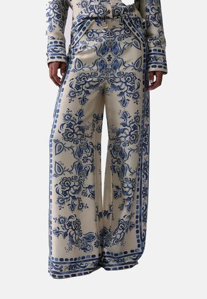 Pantaloni a gamba larga color crema con motivi floreali e paisley blu intricati, abbinati a una maglia a maniche lunghe coordinata, indossati da una persona in piedi.