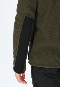 Giacca in pile verde oliva con una superficie texturizzata, maniche in soft shell nere e polsini aderenti. Dettagli delle cuciture visibili lungo la spalla.