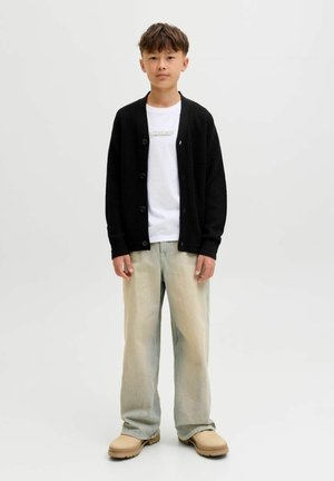 Zwarte geribbelde cardigan over een witte t-shirt met logo, gecombineerd met lichtblauwe vervaagde wijde jeans en beige schoenen.