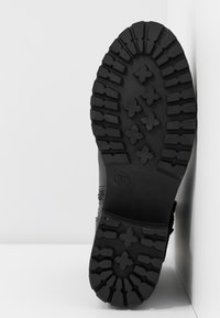 Svart gummistövelsula med djupa mönster, med geometriska mönster och en cirkulär accent. Storlek angiven som 37 på sidan.