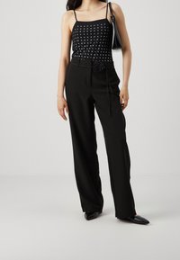 Kobieta ubrana w czarną bezrękawnik z ćwiekami, czarne spodnie z wysokim stanem i kwiatowym pasem, czarne szpilki oraz trzymająca czarną torebkę.