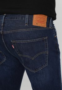 Tummansiniset farkut klassisella suoranlahjeisella leikkauksella, joissa on takataskut ja vaaleanruskea nahkapatch, jossa lukee "Levi Strauss & Co."