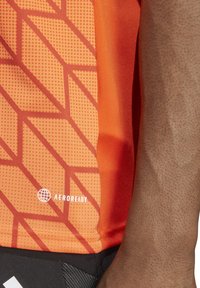 Débardeur de sport orange avec un motif géométrique, comportant des accents en points et un logo "AEROREADY". Fabriqué en matériau lisse et léger.