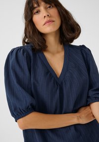 Blusa azul marino con escote en V y mangas cortas abullonadas, con un patrón de rayas texturizadas y detalles fruncidos en los puños de las mangas.
