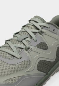 Primer plano de un zapato deportivo gris con tejido de malla, superposiciones de plástico y cordones atados sobre un fondo blanco.