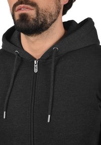 Sort zip-up hættejakke med snore, der har en glat struktur og metallisk lynlåsdetalje. Nærbillede af brystet og halsudskæringen.