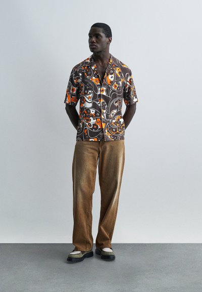 Άνδρας που φορά καφέ παντελόνι, δίχρωμα μπεζ και πράσινα παπούτσια και κοντομάνικο καφέ πουκάμισο με λαμπερά πορτοκαλί ανθικά μοτίβα, στέκεται μέσα σε εσωτερικό χώρο.