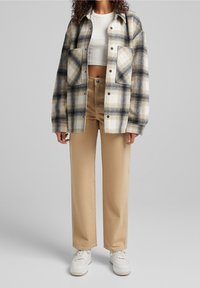 Mujer con pantalones rectos beige, top corto blanco, camisa de cuadros oversized en beige y negro, y zapatillas blancas frente a un fondo gris.