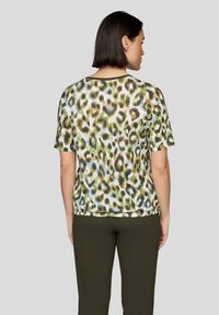 Frau mit kurzem dunklem Haar, die ein kurzärmeliges Tiermusterhemd und dunkle Hosen trägt, von hinten vor einem schlichten grauen Hintergrund stehend.