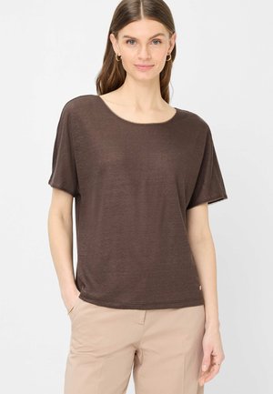 Femme aux cheveux bruns et raides portant un haut ample à manches courtes marron foncé et un pantalon beige, debout avec une main dans la poche.