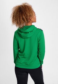 Groene hoodie met een gestructureerde afwerking en zwarte accentdetails op de schouders, gedragen met zwarte broek met zijstrepen.