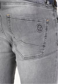 Jeans de denim gris con un bolsillo trasero bordado con un logo en relieve, detalles de costura en naranja y una textura suave, ligeramente desgastada.