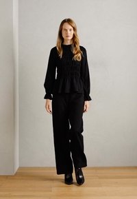 Schwarzes Raffung-Peplum-Top und weite Hosen, beide aus einem strukturierten Stoff, kombiniert mit eleganten schwarzen Stiefeletten. Neutraler Hintergrund.