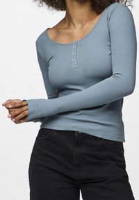 Kvinna i en figurnära blå ribbad långärmad tröja med knappar och svarta byxor, med händerna ihopknäppta.