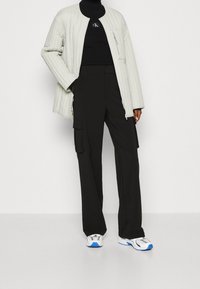 Gequilted lichtgrijze jas met een ronde hals, zwarte aansluitende coltrui, zwarte cargobroek en witte sneakers met blauwe accenten.